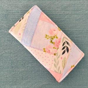 May include: A handmade patchwork wallet featuring a floral rose pattern, polka dots, and leaf designs in pink, blue, and white. The wallet is rectangular with a stitched border and a soft, quilted appearance.