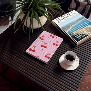 Puede incluir: Un cuaderno rosa con un estampado de cerezas, con cerezas rojas con lazos y pequeños corazones sobre un fondo a rayas. Una taza de café y una revista de viajes también están sobre la mesa de madera oscura.
