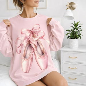 May include: A light pink long-sleeve sweatshirt featuring a large graphic print of two ballet slippers with long, flowing satin ribbons tied in a bow at the top. The slippers are rendered in shades of soft pink with subtle highlights and shadows.