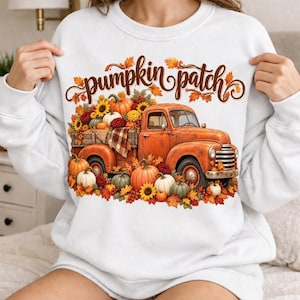 Puede incluir: Una sudadera blanca de manga larga con un estampado gráfico de un camión vintage naranja cargado de calabazas y hojas de otoño. El texto "pumpkin patch" está escrito en una fuente cursiva marrón sobre el camión, rodeado de follaje otoñal.