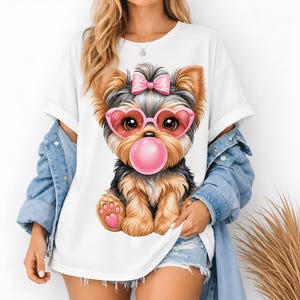 Puede incluir: Una camiseta blanca con una ilustración gráfica de un cachorro de Yorkshire Terrier de pelaje marrón y negro. El cachorro lleva gafas rosas en forma de corazón, un lazo rosa de lunares en la cabeza y sopla una gran burbuja de chicle rosa.