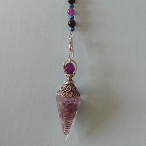 May include: A silver-toned metal chain pendulum featuring a crescent moon charm at the top. The chain is adorned with small purple and dark beads. The pendulum terminates in a faceted, conical purple stone encased in an ornate silver metal cap.