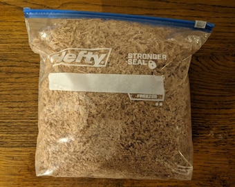 May include: A clear plastic freezer bag filled with brown wood shavings. The bag has a blue zipper closure and the word "Hefty" printed on the front. The bag also has a white label for writing and the words "Stronger Seal" and "Freezer" printed on it.