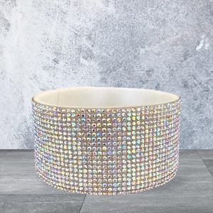 May include: A wide, cylindrical bangle bracelet featuring a dense, uniform grid of iridescent, multi-faceted rhinestones that reflect light in various colors. The interior of the bracelet is a smooth, solid off-white material with a visible seam.