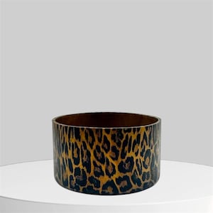 May include: A cylindrical container with a leopard print pattern in shades of brown and black. The container has a dark brown interior and sits on a white surface.