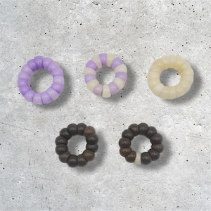 May include: Five spiral hair ties in various colors. The colors include light purple, a mix of purple and white, cream, and two shades of brown. The hair ties are made of a flexible, beaded material and are arranged on a textured gray surface.