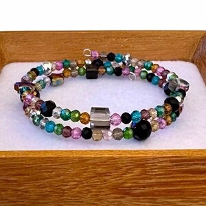 May include: A multi-strand wrap bracelet featuring a variety of faceted glass beads in shades of black, teal, emerald green, purple, and metallic silver. The design includes a central clear gray cube bead and a larger round black bead.