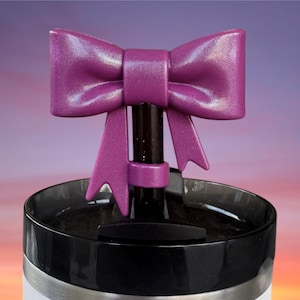 May include: A decorative bottle stopper featuring a shimmering purple bow design. The bow is mounted on a black stem and sits atop a black and silver container. The background has a gradient of purple, orange, and yellow.