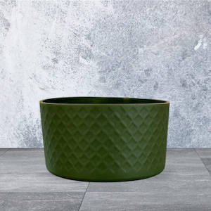 May include: An olive green, oval-shaped planter with a diamond pattern texture. The planter is made of a solid material and sits on a gray tiled surface. The background is a textured gray wall.