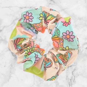 May include: A colorful scrunchie with a floral and butterfly pattern. The fabric features a mix of pastel colors, including light blue, pink, orange, and green. The design includes flowers and butterflies.