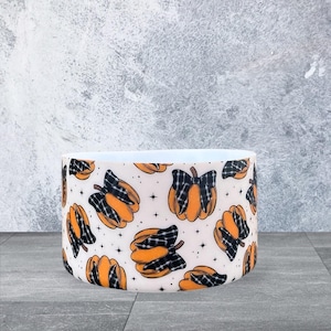 May include: A cylindrical tumbler boot featuring a repeating pattern of orange pumpkins adorned with black and white plaid bows, accented by small black stars on a white background. The design covers the exterior surface of the accessory.