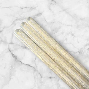 May include: Three clear, cylindrical craft sticks with gold glitter suspended within. The sticks are arranged diagonally against a white and gray marble background. The glitter catches the light, creating a sparkling effect.