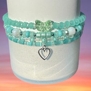May include: Three beaded bracelets in shades of turquoise and clear, stacked on a white cylindrical form. The top bracelet has round beads, the middle has a butterfly charm, and the bottom has a heart charm.