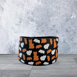 May include: A black cylindrical storage container with a repeating pattern of orange and white bear-like figures. The container is set against a gray background and floor.