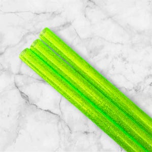 May include: A close-up of several neon green, glitter-infused straws. The straws are cylindrical and appear to be made of a clear material. They are arranged diagonally against a white and gray marble background.