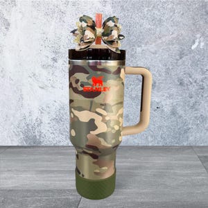 Pu&ograve; includere: Un bicchiere Stanley con motivo mimetico, manico beige e base verde. Il bicchiere presenta il logo Stanley in rosso ed &egrave; sormontato da un fiocco decorativo. Progettato per mantenere le bevande calde o fredde.
