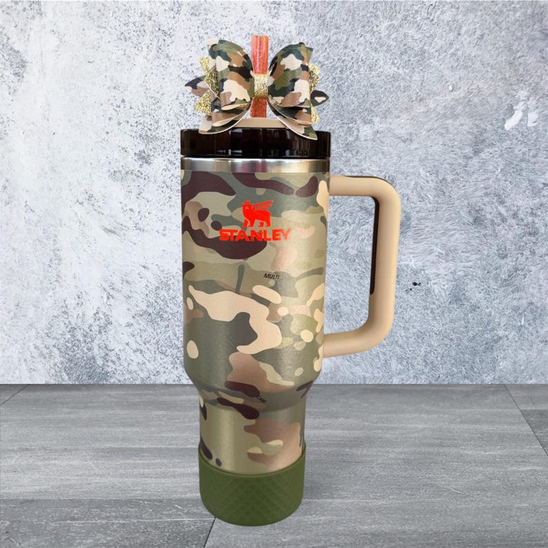 Pu&ograve; includere: Un bicchiere Stanley con motivo mimetico, manico beige e base verde. Il bicchiere presenta il logo Stanley in rosso ed &egrave; sormontato da un fiocco decorativo. Progettato per mantenere le bevande calde o fredde.