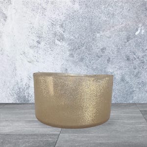 May include: A gold-colored, glitter-covered, oval-shaped container. The container has a smooth, rounded base and a wide opening. The background is a textured gray and white wall, and the container sits on a gray tiled surface.