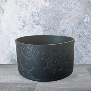 May include: A matte black cylindrical planter featuring a debossed spider web pattern across its exterior surface. The container has a wide, open top and a flat base, providing a deep interior space for holding various items or small plants.