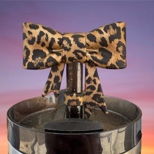 May include: A decorative bow tie with a leopard print pattern in brown and black, mounted on a dark, cylindrical stand. The stand is set on a black container with a silver band, against a gradient background.