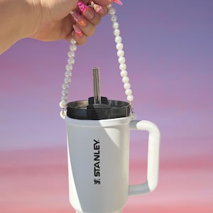 May include: A white Stanley tumbler with a black lid and a metal straw, featuring a decorative carrying strap made of white faux pearls attached to the sides. The brand name Stanley is printed vertically in black text on the side of the cup.