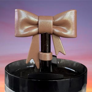 May include: A brown bow-shaped bottle stopper with a metallic sheen, attached to a black and clear container. The bow has detailed folds and a decorative design. The background has a gradient of pink, purple, and orange.