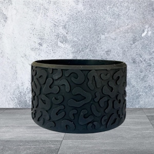 May include: A black, cylindrical drink sleeve with a textured, abstract pattern. The sleeve is made of a flexible material and is designed to fit around a standard-sized beverage container. The background is a gray, textured wall and a gray floor.