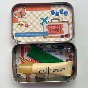 Può includere: Una scatola di metallo rettangolare aperta con decorazioni a tema viaggio. All'interno, un tubetto di balsamo per le labbra, tagliaunghie e altri piccoli oggetti sono visibili. Il coperchio presenta una farfalla, un aereo e un biglietto con la scritta "City Bus". Il testo dice "you can't buy happiness but you can go to Spain."