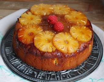 Pode incluir: Um bolo invertido de abacaxi redondo em um prato decorativo. O bolo é marrom dourado com fatias de abacaxi caramelizadas e duas framboesas vermelhas no topo. O prato é escuro com uma borda texturizada, sobre um pano branco.