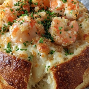 May include: A close-up shot of a bread bowl filled with cheesy shrimp scampi. The shrimp are pink and orange, topped with parsley. The bread bowl is golden brown with a soft interior, and a piece is broken off.