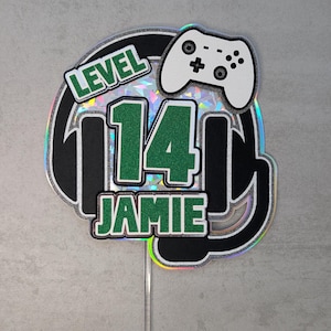 May include: A cake topper with a gaming theme. The design features a black and silver headset shape, with the words "LEVEL 14 JAMIE" in green and white. A white game controller is at the top.