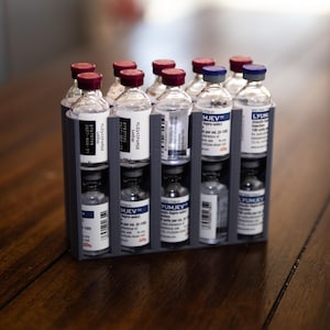 May include: A gray plastic organizer holding multiple small glass vials with red and blue caps. The vials have white labels with black text. The organizer is on a wooden surface.