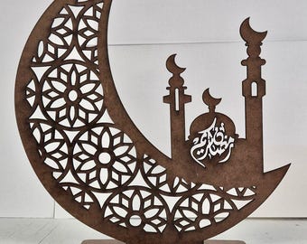 May include: A brown wooden crescent moon and mosque silhouette. The moon features a decorative cutout pattern. The mosque has two minarets and a dome, with Arabic calligraphy in the center. A decorative item for Ramadan.