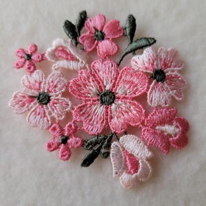 以下が含まれることがあります： ピンクと白の色合いで刺繍された花のクラスターが特徴の繊細な花のアップリケ。濃い緑色の葉が付いています。花は詳細なステッチと中央の濃い灰色のアクセントがあり、衣服や工芸品を飾るのに最適です。