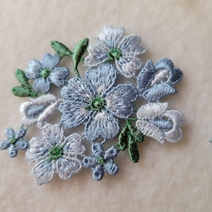 以下が含まれることがあります： 青と白の色合いに緑のアクセントが施された刺繍の花柄アップリケ。 花には詳細なステッチが施され、クラスター状に配置されています。 衣類やクラフトプロジェクトの装飾に最適です。
