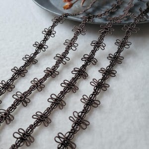 以下が含まれることがあります： 濃い茶色の装飾用ワイヤートリムの4本のストランドのクローズアップ。各ストランドは、様式化された花またはリボンのような形状の繰り返しパターンを特徴としています。トリムは明るい背景に対して斜めに配置されており、工芸品やジュエリーの供給を示唆しています。