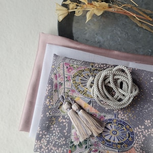 以下が含まれることがあります： テキスタイルと装飾要素の詰め合わせ。花柄のグレーの生地は、白い生地、ブラッシュピンクのシルク、タッセル付きのシルバーコードと組み合わされています。背景にはドライフラワーのアクセントがあります。