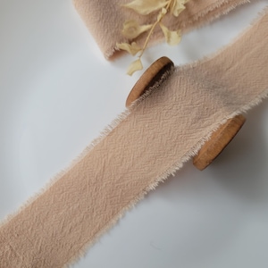 以下が含まれることがあります： ベージュ色の布リボン。縁はほつれており、木製の糸巻きから部分的にほどかれています。リボンは質感のある素材でできています。乾燥した花がリボンに置かれ、自然な美しさを演出しています。