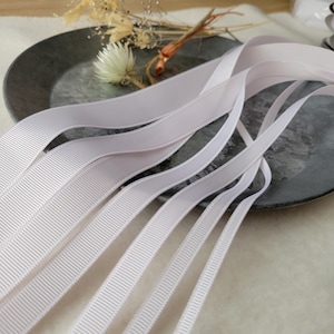 以下が含まれることがあります： さまざまな幅の白いグログランリボンが、濃い灰色の金属トレイに配置されています。リボンはリブ編みのテクスチャで、わずかにカーブしています。背景にはドライフラワーとグリーンが見えます。