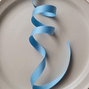 以下が含まれることがあります： ニュートラルグレーの背景に、優雅に渦巻く水色のリボン。リボンは滑らかでサテンのような質感で、クラフト、ギフト包装、または装飾目的で使用される可能性があります。リボンの色はソフトなパステルブルーです。