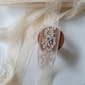 以下が含まれることがあります： アイボリー色のレースリボン、花柄のデザイン。繊細なレースは木製のスプールに部分的に巻き付けられています。リボンは柔らかいオフホワイト色で、透け感のある素材で作られているようです。