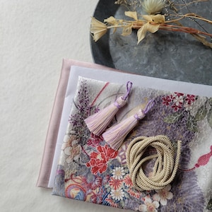 以下が含まれることがあります： 赤と白の花柄の生地、2つのラベンダー色のタッセル、丸まった金色のコードなど、さまざまなクラフト用品。淡いピンク色の生地と、ドライフラワーが入った金属製のトレイも写っています。