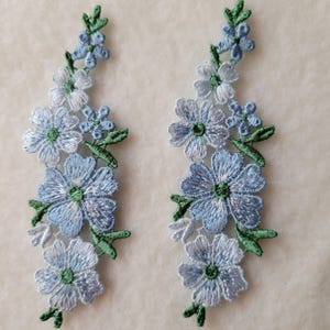 以下が含まれることがあります： 2つの刺繍された花のアップリケ。各アップリケは、緑の葉を持つ青と白の花の茎を特徴としています。花は、より濃い青色の中心部を持つ、詳細で質感のある外観をしています。
