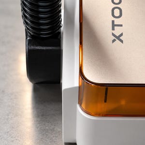 May include: A white and gold XTOOL device featuring a black corrugated exhaust hose attached to the side. The unit has a translucent amber-colored plastic panel along the edge and a smooth white base, with the brand name XTOOL printed vertically.