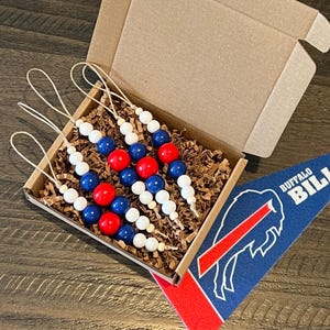 May include: A set of four beaded ornaments in a cardboard box with shredded paper. The ornaments feature alternating red, white, and blue beads. A blue and red pennant with the words "Buffalo Bills" is also in the image.