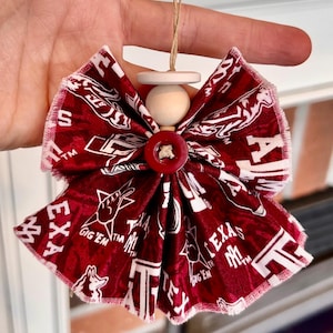 Peut inclure: Ornement en tissu bordeaux et blanc en forme de nœud papillon. Le tissu présente les inscriptions "TEXAS A&M" et "GIG'EM", avec un bouton central. Une perle et un disque en bois sont en haut, attachés avec de la ficelle.