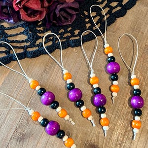 May include: Six beaded ornaments with a Halloween theme. Each ornament has a string loop and features a large purple bead, black beads, orange beads, and white beads. The ornaments are on a wooden surface.