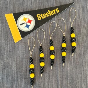 May include: A black and gold pennant with the word "Steelers" and a team logo, along with five beaded ornaments in black and yellow. The ornaments have a loop of twine for hanging. The background is a gray fabric.
