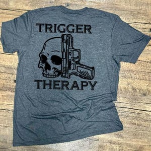May include: A gray t-shirt with the words "TRIGGER" and "THERAPY" printed above and below a black graphic of a skull and a handgun. The shirt is made of a soft material.