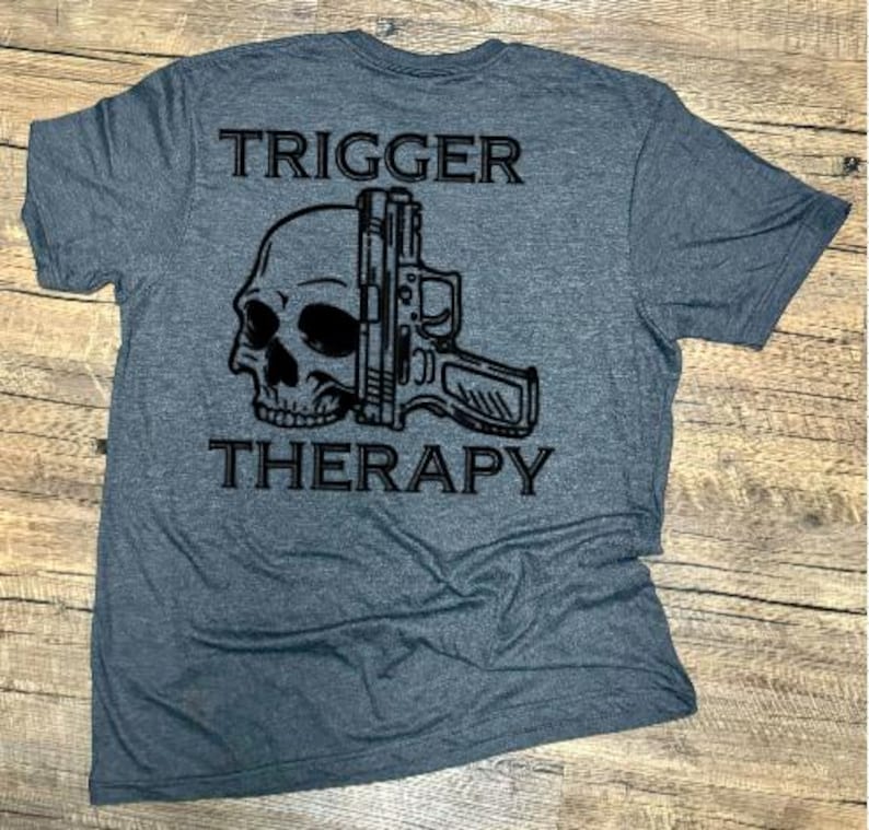 May include: A gray t-shirt with the words "TRIGGER" and "THERAPY" printed above and below a black graphic of a skull and a handgun. The shirt is made of a soft material.
